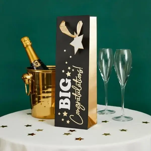 Black and gold gift bag with Big Congratulations text beside a champagne bucket and two flutes on a decorated table.